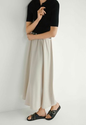 Osoba ubrana w czarną bluzkę z krótkim rękawem, beżową midi spódnicę z wysokim stanem i paskiem oraz czarne klapki z ćwiekami, stojąca na tle białej ściany.