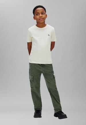 Ragazzo in piedi con le mani dietro la schiena, indossa una t-shirt crema a maniche corte e pantaloni cargo verdi su uno sfondo grigio uniforme.