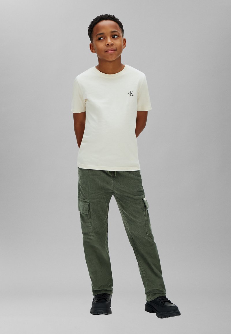 Garçon debout, les mains dans le dos, portant un t-shirt crème à manches courtes et un pantalon cargo vert sur un fond gris uni.