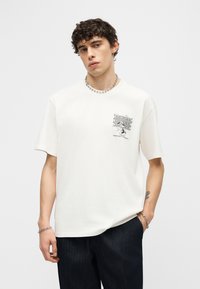 Νεαρός άνδρας με σγουρά μαλλιά φοράει λευκό μπλουζάκι με γραφικό σχέδιο και σκούρο παντελόνι, διακοσμημένος με αλυσίδα στον λαιμό, βραχιόλι και σκουλαρίκι, στέκεται μπροστά σε λευκό φόντο.