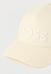 Kremna baseball kapa s strukturiranim dizajnom, z vvezanim logotipom "BOSS" v beli barvi, teksturirano tkanino in ukrivljenim ščitnikom.