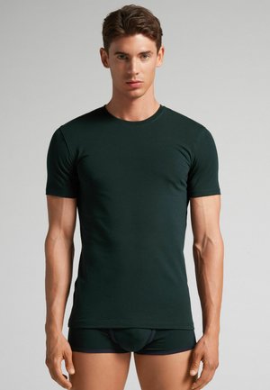 Dunkelgrünes T-Shirt mit kurzen Ärmeln aus einem weichen, dehnbaren Stoff, mit einem Rundhalsausschnitt und einer taillierten Passform, kombiniert mit schwarzen Shorts.