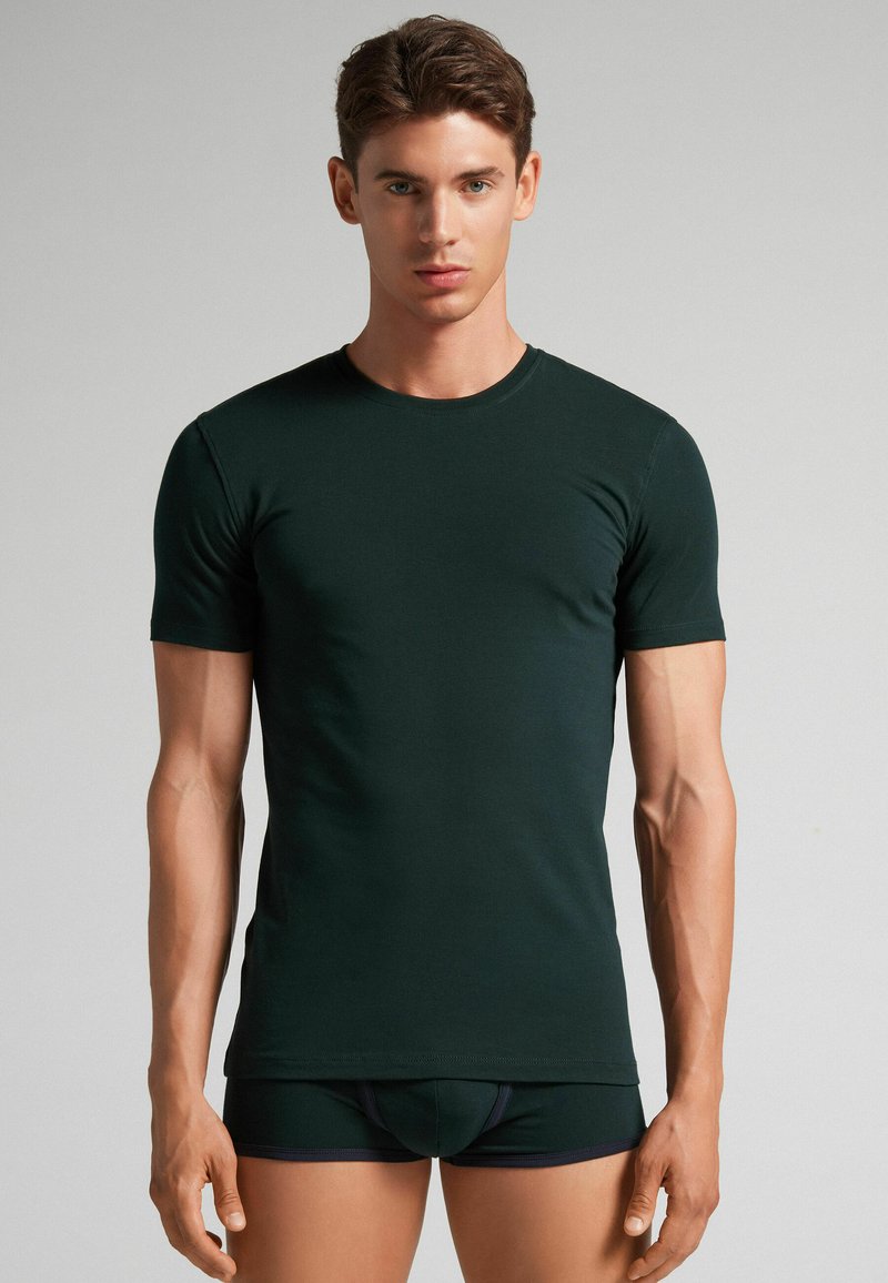 Dunkelgrünes T-Shirt mit kurzen Ärmeln aus einem weichen, dehnbaren Stoff, mit einem Rundhalsausschnitt und einer taillierten Passform, kombiniert mit schwarzen Shorts.