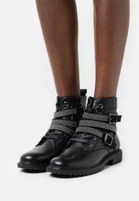 Botas de tobillo de cuero negro con correas de tachuelas plateadas, cierre de cordones en la parte delantera, cremallera lateral y suela de goma texturizada.