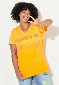 T-shirt en coton orange vif avec un col en V, arborant un texte scintillant "HEUREUX EN ORANGE". Assorti avec un jean bleu, affichant une coupe décontractée et relaxée.