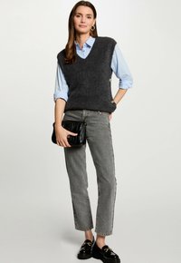 Maglione grigio a maglia con scollo a V, camicia blu sotto, jeans grigi dritti e una borsa a tracolla nera trapuntata. Mocassini neri completano il look.