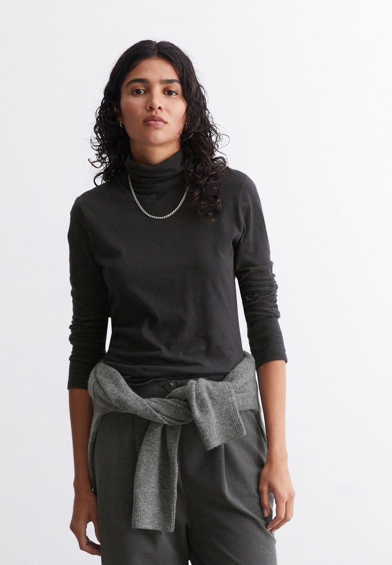 Maglietta a maniche lunghe nera con collo alto, in tessuto morbido. Maglione grigio annodato in vita, pantaloni grigi a sigaretta, modella con capelli ricci, sfondo neutro.