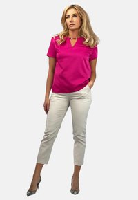 Fuksjowa bluzka krótki rękaw z dekoltem w serek i metalicznym akcentem, zestawiona z jasnobeżowymi spodniami wąskimi i wzorzystymi szpilkami.