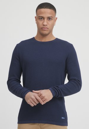 Marineblå genser med lange ermer, strukturert strikket stoff, rund hals, tettsittende mansjetter, og en liten merkeetikett nederst.