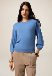 Pull en maille bleu clair avec un décolleté large et des manches trois-quarts, associé à un pantalon beige. Texture lisse et coupe décontractée.