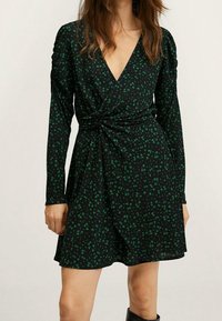 Robe noire cintrée ornée de motifs floraux verts. Elle possède des manches longues et un décolleté en V profond. Le tissu tombe doucement.