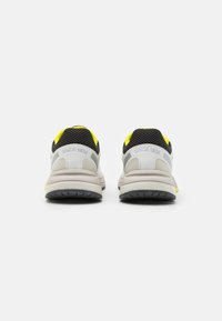 Sneakers con una toma in rete grigia, accenti neri e dettagli gialli sul tallone. Suola intermedia bianca spessa e suola esterna nera texturizzata.