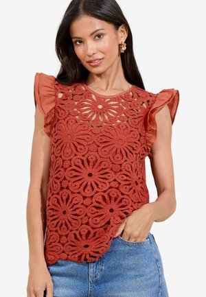 Top color ruggine con motivo in pizzo floreale, maniche arricciate e scollo rotondo, abbinato a pantaloni shorts di jeans. Dettagli del tessuto testurizzato visibili.