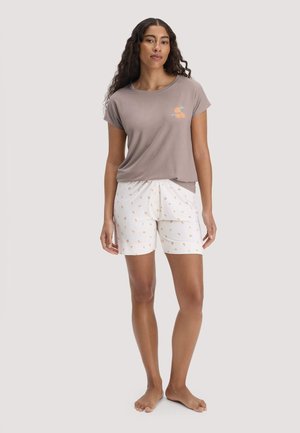 Frau trägt ein taupefarbenes kurzärmeliges Shirt mit orangefarbenem Grafikmuster und weiße Shorts mit kleinen orangefarbenen Fruchtmustern, steht barfuß.