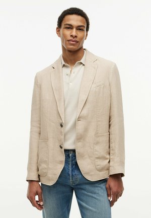 Ung mand iført en beige blazer over en lys polo-shirt og blå jeans, stående med en afslappet holdning mod en ensfarvet hvid baggrund.