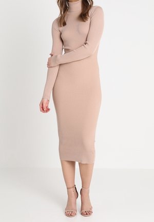 Femme portant une robe midi beige à col roulé côtelée à manches longues, associée à des talons ouverts à brides de cheville assortis, debout devant un fond uni.