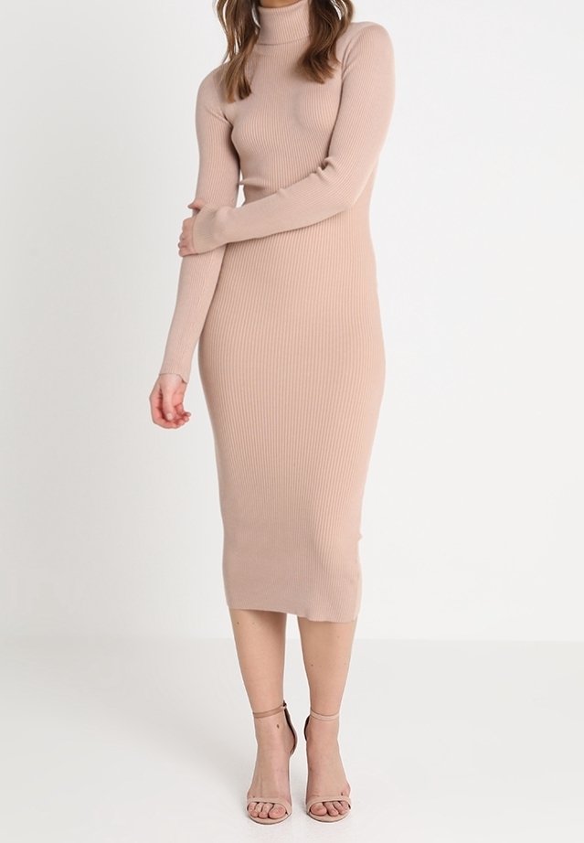 Femme portant une robe midi beige à col roulé côtelée à manches longues, associée à des talons ouverts à brides de cheville assortis, debout devant un fond uni.