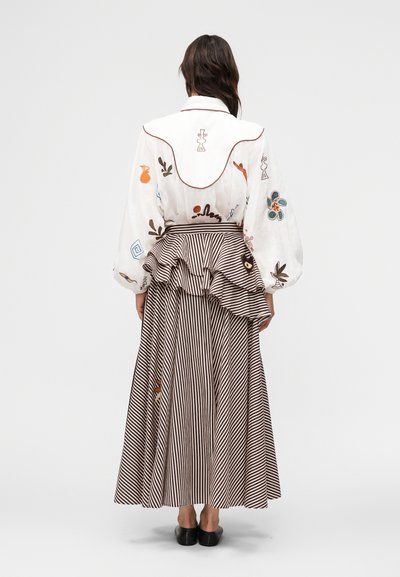 Γυναίκα γυρισμένη πλάτη, φορούν λευκό πουκάμισο με πολύχρωμη κεντητή διακόσμηση και φούστα με καφέ-λευκές ρίγες και βολάν, με μαύρα επίπεδα παπούτσια.