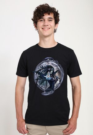 Jeune homme souriant, portant un T-shirt noir avec une illustration gothique détaillée d'une fée tenant un objet lumineux, pantalon beige, fond clair uni.