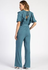 Teal jumpsuit met gerimpelde mouwen en een open rugdetail, met een knoopsluiting. Uitlopende pijpen en een gladde stoftextuur.
