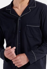 Maglietta da pigiama in cotone blu navy con colletto corto, piping bianco lungo i bordi, taschino anteriore e chiusura con bottoni. Presenta maniche lunghe e una vestibilità rilassata.