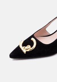 Chaussure en velours noir à bout pointu avec une grande boucle décorative en forme de nœud doré sur le dessus avant.