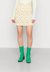 Saia mini branca com padrão floral amarelo e barras franzidas, combinada com botas de tornozelo verdes brilhantes com salto quadrado.