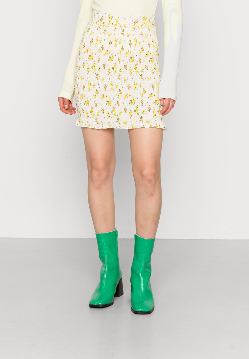 Saia mini branca com padrão floral amarelo e barras franzidas, combinada com botas de tornozelo verdes brilhantes com salto quadrado.