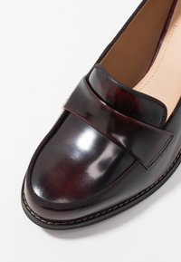 Mocassin en cuir verni bordeaux avec un large nœud lisse en accent et des coutures contrastantes le long de la semelle, présentant un design à bout rond.