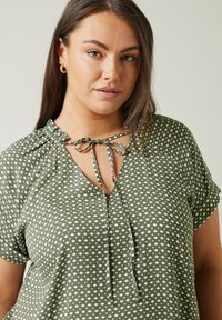 Grüne Bluse mit Polka-Dots, V-Ausschnitt, Bindedetail am Ausschnitt und kurzen Ärmeln. Der Stoff hat eine weiche Textur und eine lockere Passform.