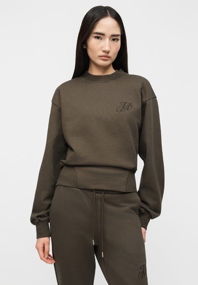 Νεαρή γυναίκα με ίσια σκούρα μαλλιά φοράει φούτερ σε ελιά πράσινο και αντίστοιχο παντελόνι φόρμας με διακριτικό κεντητό λογότυπο στο στήθος και στο πόδι.