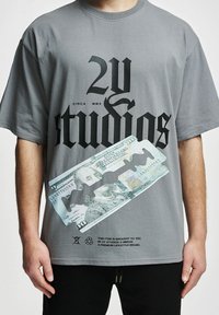 Camiseta de algodón gris con texto negro en negrita, un gráfico de un billete de $100 y un diseño de una hoja de afeitar. La etiqueta adjunta indica la marca y las instrucciones de cuidado.