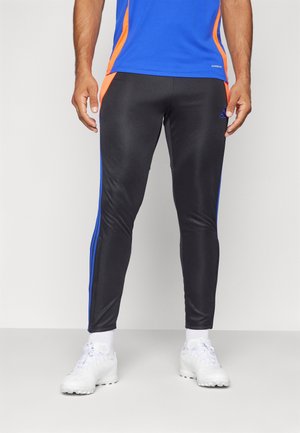 Homme portant un pantalon de sport noir avec des bandes bleues, des accents orange et des baskets blanches, debout devant un fond clair uni.
