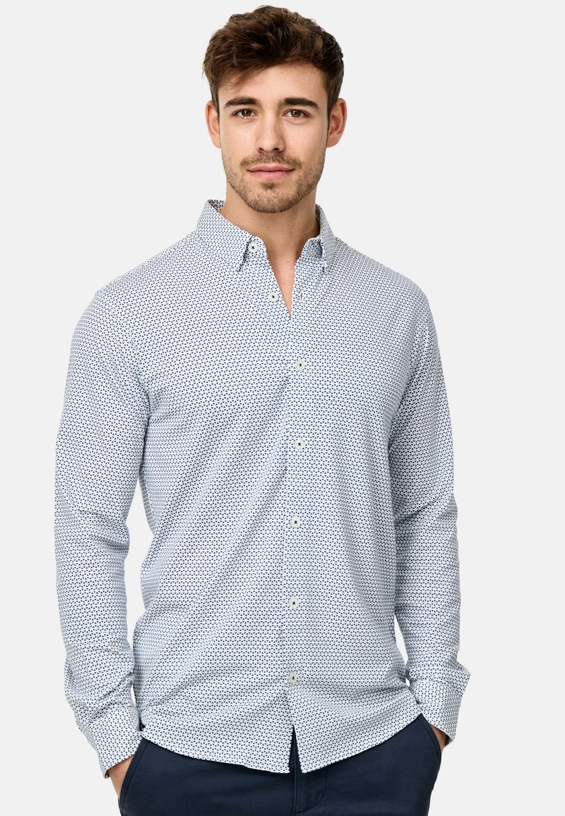 Camicia bianca con un motivo a pois blu, maniche lunghe e colletto classico. Il tessuto sembra liscio con un design attillato.