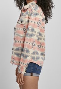 Button-up shirt featuring a geometric pattern in pink, blue, and cream. Long sleeves, collar, and two chest pockets. Denim shorts visible below.