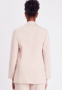 Blazer rose clair avec une coupe ajustée, manches longues et une fente au dos. Texture lisse et détails matériels minimaux.