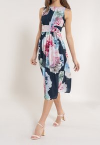 Robe à imprimé floral bleu marine foncé avec de grandes fleurs roses et bleues. Sans manches, avec un corsage plissé et une jupe fluide. Associée à des talons roses clair.