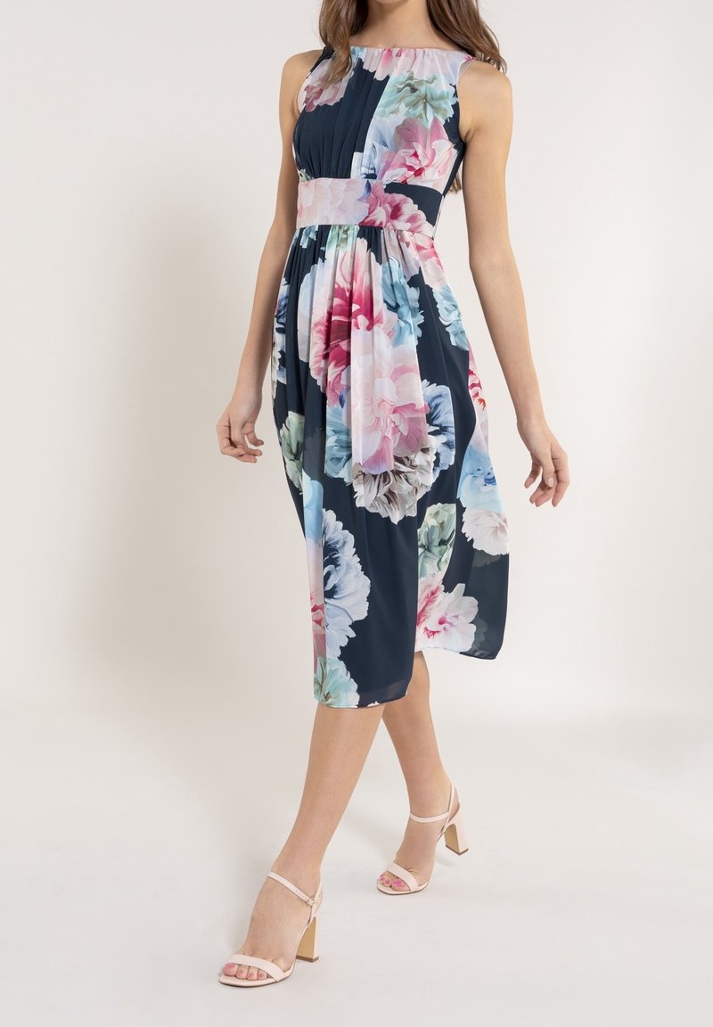 Robe à imprimé floral bleu marine foncé avec de grandes fleurs roses et bleues. Sans manches, avec un corsage plissé et une jupe fluide. Associée à des talons roses clair.