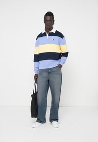 Maglietta da rugby a maniche lunghe in blu chiaro, con righe gialle e blu navy; colletto bianco e logo; abbinata a jeans blu larghi e una borsa a tracolla nera.