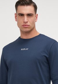 Tricou cu mânecă lungă de culoare bleumarin, din bumbac, cu gât rotund, având un logo alb "REPLAY" cusut pe piept. Textură netedă.