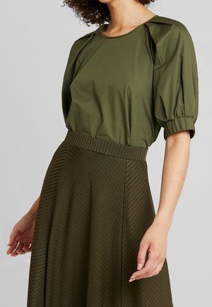 Kobieta ubrana w oliwkowo-zieloną bluzkę z bufkami i dopasowaną, wysokociśnieniową, teksturowaną spódnicę na jednolitym tle.