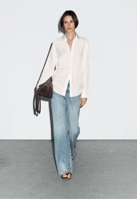 White button-up shirt with long sleeves, paired with light blue wide-leg jeans and a textured brown handbag. Model wears flat sandals.