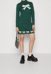 Grön stickad klänning med hög krage, vit rosettdetalj och prickar. Har en halsad fåll med rosettmönster. Parat med svarta platåskor.