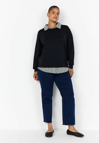 Maglia nera a maniche lunghe con una camicia a righe colletto sotto, abbinata a jeans blu e ballerine nere intrecciate; look informale con texture a contrasto.