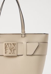 Borsa tote beige realizzata in materiale strutturato, con una grande placca logo, manici lunghi e accenti laterali angolari.