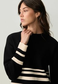 Pull noir avec des rayures crème aux poignets et à la taille, doté d'une texture côtelée et d'une coupe décontractée. Design minimaliste, col rond.