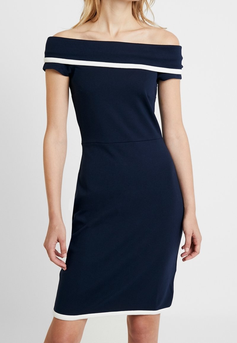 Femme portant une robe bleu marine ajustée, épaules dénudées, avec un liseré blanc le long de l'encolure et de l'ourlet sur un fond uni.