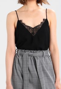 Femme portant un caraco noir à bretelles fines orné de dentelle, rentré dans une jupe taille haute à motif écossais noir et blanc avec ceinture.