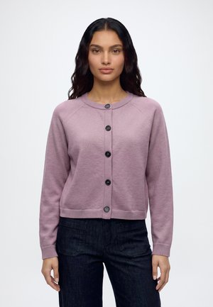 Femme aux longs cheveux foncés portant un cardigan boutonné violet clair et un pantalon foncé, faisant face à un fond clair uni.