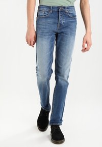 Homme portant un jean slim bleu délavé et des chaussures noires à lacets, marchant devant un fond blanc uni.